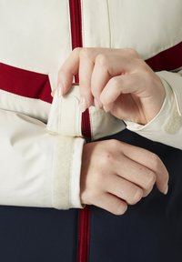 White jacket with red accents, featuring a zip closure and adjustable Velcro cuff. Smooth exterior with a soft, textured inner cuff.