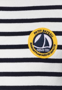 Gestreifter Stoff in Marineblau und Weiß mit einem runden gestickten Patch, das ein Segelboot-Design und den Text "PETIT BATEAU depuis 1893" zeigt.