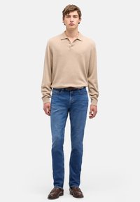 Beżowa koszulka polo z długim rękawem i trzema guzikami, w zestawieniu z niebieskimi dopasowanymi dżinsami i brązowymi eleganckimi butami. Gładka dzianinowa tekstura na koszulce.