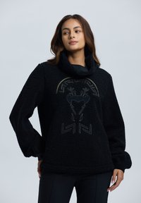 Czarny polarowy sweter z wysokim kołnierzem i ściągaczami. Posiada złoty graficzny nadruk głowy jelenia z dekoracyjnymi wzorami. Miękka tekstura.