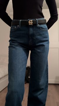 Blå denimjeans med løs pasform, der har et sort bælte og en gylden dobbelt "B" spænde. Teksturen er glat med minimal falmen.