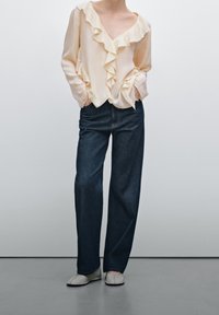 Blusa beige chiaro con fronte a volant, abbinata a jeans a gamba larga blu scuro e scarpe slip-on chiare. Tessuto morbido, vestibilità casual.