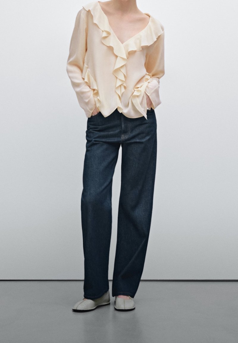 Blusa beige chiaro con fronte a volant, abbinata a jeans a gamba larga blu scuro e scarpe slip-on chiare. Tessuto morbido, vestibilità casual.