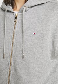 Sweat à capuche zippé gris en tissu doux, avec une fermeture éclair dorée, un logo contrastant sur la poitrine gauche et des cordons avec embouts en métal.