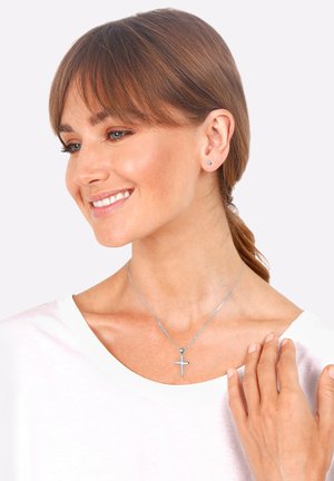 Silberne Kreuzkette an einer zarten Kette, getragen mit einem schlichten weißen Oberteil. Kleine Ohrstecker ergänzen den Look. Sanfte Texturen, unauffälliges Design.