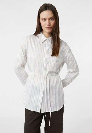 White button-up shirt with thin vertical stripes in beige, featuring a collar and a tied waist. Long sleeves with a slightly curved hem.