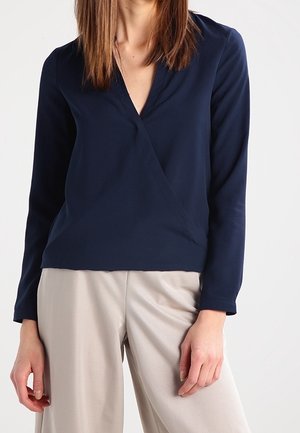 Marineblå omslagstop med lange ærmer, V-udskæring og glat stof. Matchet med lys beige vide bukser. Enkel design og glat tekstur.