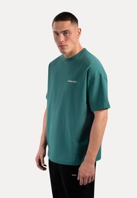 Camiseta de manga corta en color verde azulado, fabricada en tejido de punto, con cuello redondo. Presenta un pequeño logo blanco en el pecho y un corte relajado.