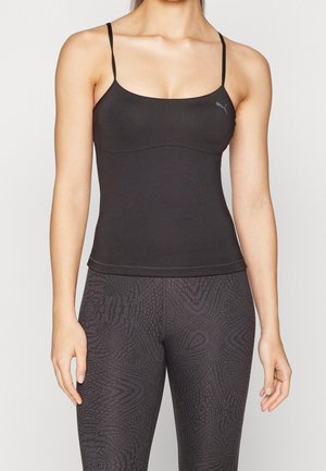 Frau trägt ein schwarzes, eng anliegendes Tanktop mit dünnen Trägern und gemusterte dunkle Leggings, steht vor einem einfarbigen hellen Hintergrund.