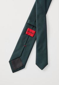 Cravate rayée vert foncé avec une pointe satinée noire et une étiquette de marque rouge. Fabriquée en Italie, avec une finition lisse et texturée.