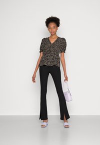 Top preto com estampado floral e mangas bufantes, calças pretas de corte justo com rachas laterais, bolsa lavanda e sapatos de salto aberto lavanda sobre um fundo neutro.