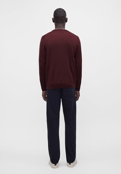 Homme debout, dos tourné vers le spectateur, portant une chemise rouge foncé à manches longues, un pantalon bleu foncé et des baskets blanches sur un fond clair uni.