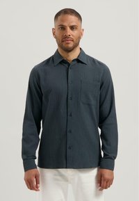 Donkerblauw gestructureerd overhemd met lange mouwen, enkele borstzak en zwarte knopen. Gepaard met witte broek.