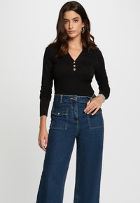 Top lungo a maniche ribattute nere con scollo a V e tre bottoni dorati. Abbinato a jeans di denim blu a vita alta e larghi, con tasche frontali.