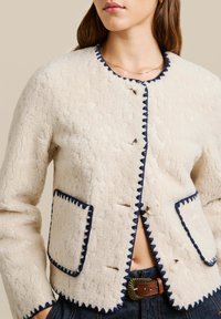 Valge fliisjakker navy ääristega, ringkaelusega, nupuga kinnitustega ning dekoratiivse tikandiga äärtes ja taskutes.