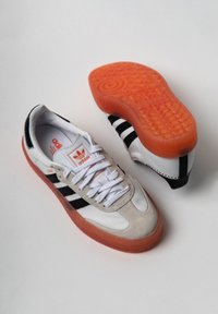 Białe i szare skórzane sneakersy z czarnymi pasek, pomarańczową gumową podeszwą oraz teksturowanymi akcentami na górze. Posiadają sznurowany krój oraz detal z logo.