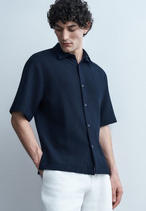 Άνδρας με σγουρά μαλλιά που φοράει μπλε πουκάμισο με κοντά μανίκια και λευκό παντελόνι, κοιτάζοντας προς τα κάτω με τα χέρια στις τσέπες, μπροστά σε ένα ανοιχτόχρωμο φόντο.