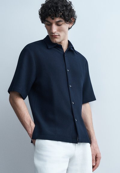 Homme aux cheveux bouclés portant une chemise bleu marine à manches courtes boutonnée et un pantalon blanc, regardant vers le bas les mains dans les poches sur un fond pâle.