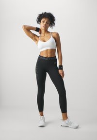 White sports bra with a branded band, paired with high-waisted black leggings featuring a vertical logo. Model wears wristband and sneakers.