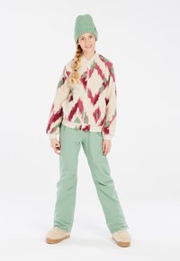 Fleece bunda s bílým, červeným a zeleným diamantovým vzorem, s předním zipem. Světle mátové voděodolné kalhoty. Texturovaná mátová čepice a huňaté pantofle.