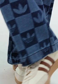Blue checkered pants with a repeating leaf pattern, paired with cream sneakers featuring brown stripes and a textured sole.