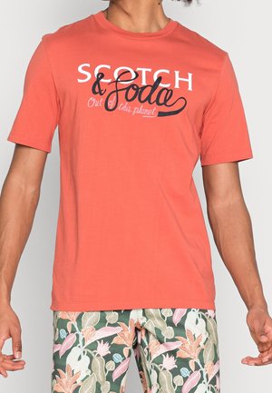 Mand iført en koralorange t-shirt med hvid og sort tekst og grønne blomstrede shorts, stående med armene afslappet ned langs siderne.