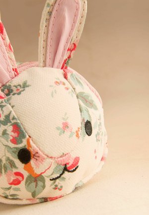 Pehmustettu kangaspupu, jossa mustat kirjotut silmät, vaaleanpunainen nenä ja kukkakuvio luonnonvalkoisella pohjalla, beigeä pintaa vasten.