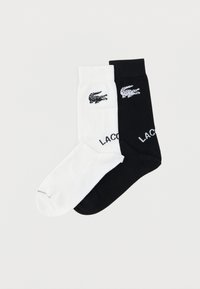 Paire de chaussettes basses, une blanche et une noire, chacune arborant un petit logo crocodile et le texte partiel "LACOSTE" près du bord-côte.