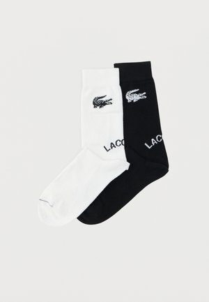 Paar Knöchelsocken, eine weiß und eine schwarz, jeweils mit einem kleinen Krokodil-Logo und teilweise sichtbarem "LACOSTE"-Schriftzug nahe der Manschette.
