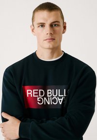 Námornícky sveter s červeným a čiernym obdĺžnikovým grafickým prvkom. Text "RED BULL RACING" v bielej farbe. Mäkko tkaný materiál, okrúhly výstrih, dlhé rukávy.