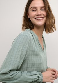 Blusa verde claro de tejido texturizado con escote en V, mangas largas y abullonadas, y un sutil patrón a cuadros. Material suave y fluido.