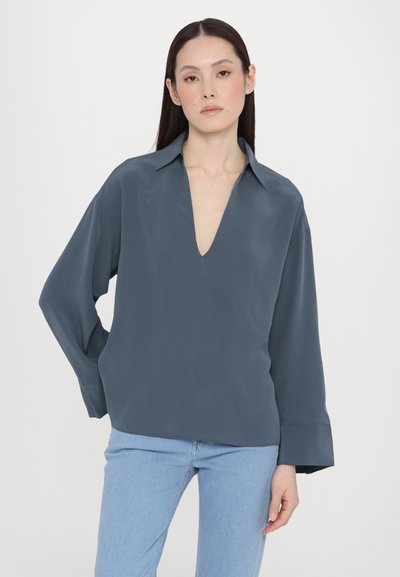 Blusa grigia a maniche lunghe con scollo a V profondo, realizzata in un tessuto morbido. Presenta una vestibilità comoda e polsini con una leggera svasatura. Abbinata a jeans blu.