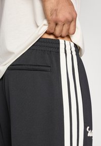 Sötétszürke sportnadrág fehér oldalsávokkal, rugalmas derékpánttal és apró zseb részlettel. Az anyag sima, enyhén fényes megjelenésű.