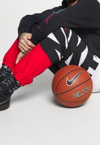 Eine Person mit schwarzen und roten Hosen sitzt auf dem Boden, hat die Hände gefaltet und lehnt sich gegen einen Nike Elite Tournament Basketball.