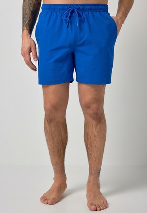 Mand iført blå svømmeshorts med snøre, står barfodet med hænderne i lommerne, viser tatoveringer på venstre arm og højre ankel.