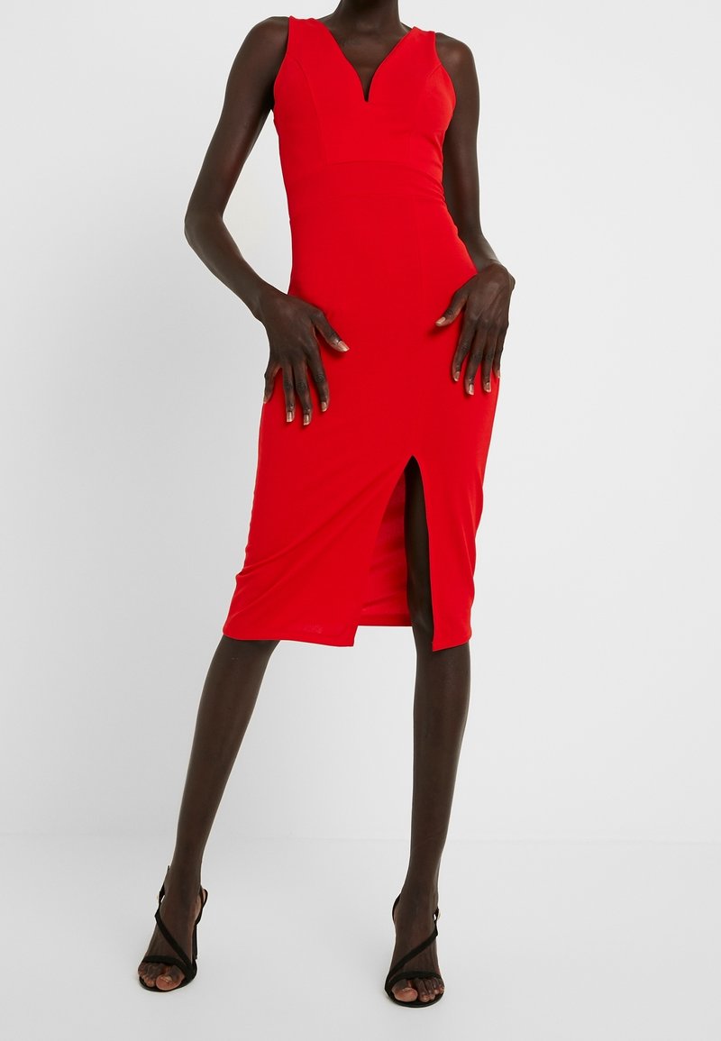 Robe rouge ajustée avec un décolleté plongeant, un ourlet à hauteur des genoux et une fente à l'avant, réalisée en tissu lisse. Portée avec des talons noirs à brides.