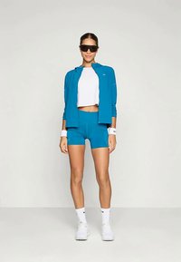 Athletic outfit featuring a turquoise zip-up jacket, white crop top, and matching shorts. Accessories include sunglasses and wristbands.