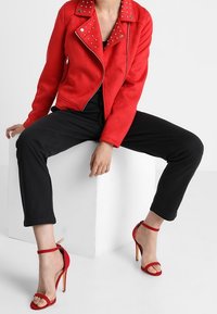 Veste de moto en simili cuir rouge avec des clous argentés sur les épaules, associée à un pantalon noir et des sandales à talons hauts rouges à lanières.