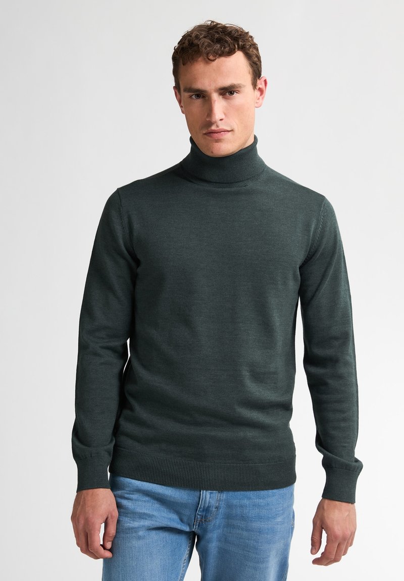 Pulover cu guler înalt de culoare verde închis, fabricat dintr-un material moale, având mâneci lungi și tiv ondulat. Asortat cu blugi din denim de culoare albastru deschis.
