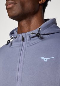 Sivi zip-up dukserica s visokim ovratnikom, podesivim šalovima i suptilnim logotipom na lijevoj strani prsa. Mekani, glatki tekstil.