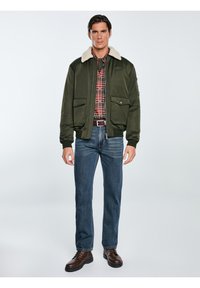 Grüne Bomberjacke mit einem beigen Kunstfellkragen, kombiniert mit einem rot-schwarzen Karoshirt, blauen Jeans und braunen Schnürstiefeln.