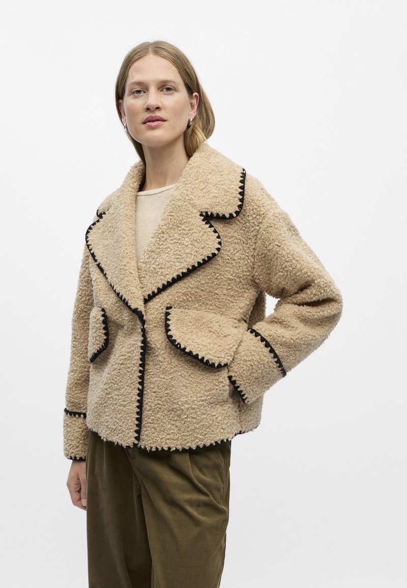 Beige Fake-Fur-Jacke mit strukturierter Oberfläche, schwarzem Kontraststich, Reverskragen und zwei großen Fronttaschen.