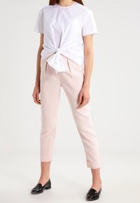 Žena v bílém tričku s krátkým rukávem uvázaném v pase, světlých růžových zkrácených kalhotách a černých mokasínách, stojící na bílém pozadí.