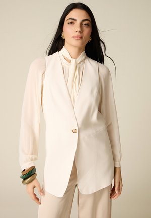 Donna con capelli lunghi e scuri indossa una camicetta color crema con fiocco al collo, blazer crema senza maniche, pantaloni beige, orecchini dorati e braccialetti spessi impilati.