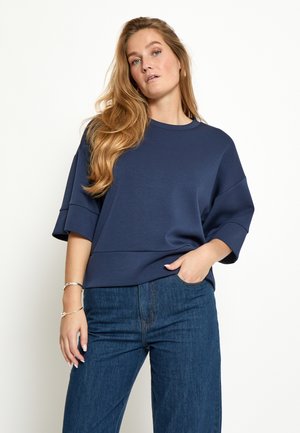 Vrouw met lang blond haar die een losse marineblauwe top en donkerblauwe spijkerbroek draagt, poserend met één hand in de zak tegen een witte achtergrond.