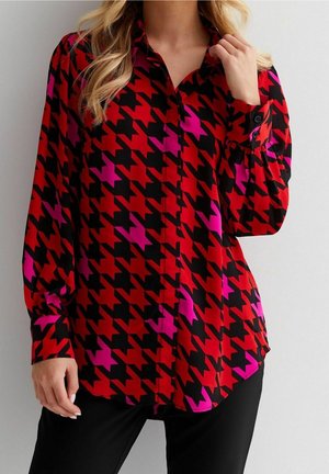 Červená, černá a růžová košile s dlouhým rukávem a vzorem loveckých zubů, nošená s černými kalhotami, zobrazena od krku k bokům.