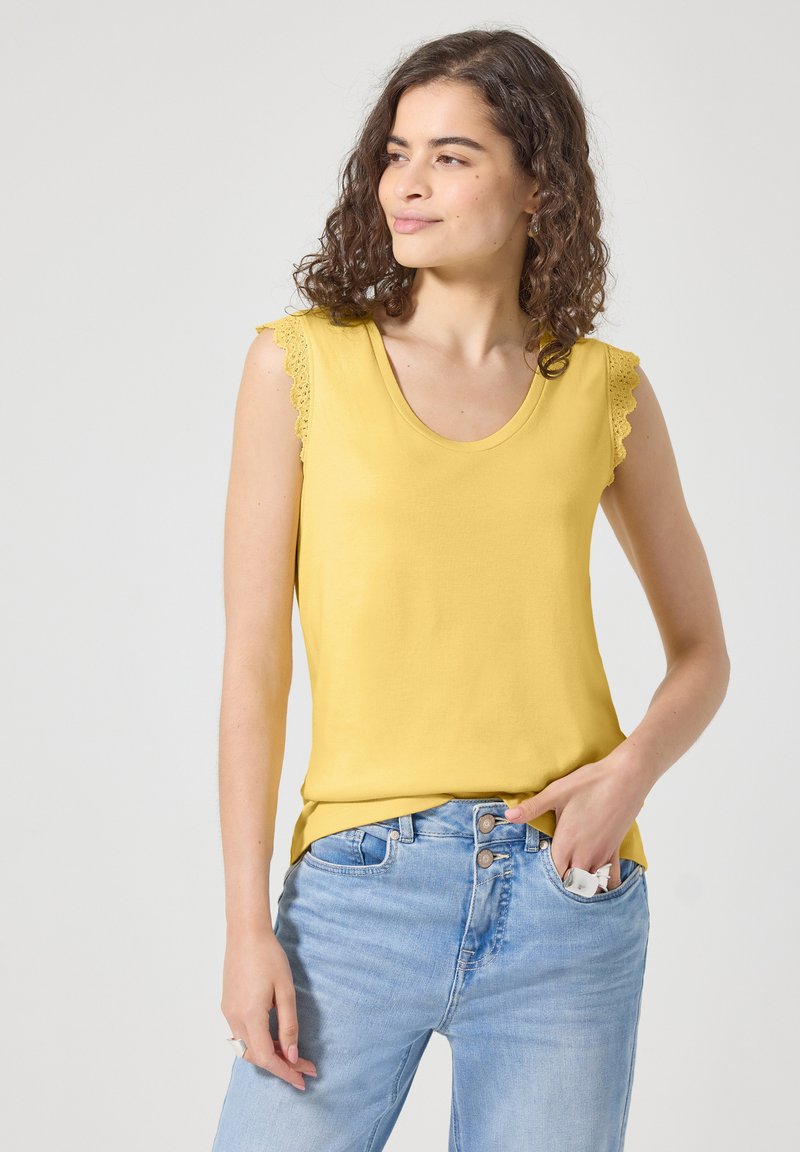 Femme aux cheveux bouclés portant un haut sans manches jaune avec un bord en dentelle et un jean bleu clair, main dans la poche tenant un objet blanc.