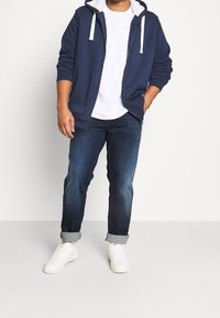 Sudadera de cremallera azul marino con cordones blancos, camiseta de cuello redondo blanca, jeans azul oscuro y zapatillas blancas. Atuendo casual con tela texturizada.