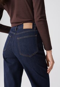 Persoon draagt donkerblauwe spijkerbroek en een bruine top met lange mouwen met de hand rustend op de heup tegen een effen lichte achtergrond.