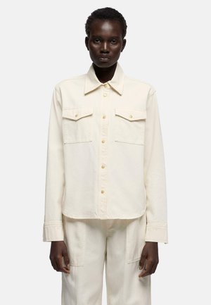 Mørkhudet person iført offwhite skjorte med knapper og brystlommer samt matchende bukser, stående mod en ensfarvet baggrund.